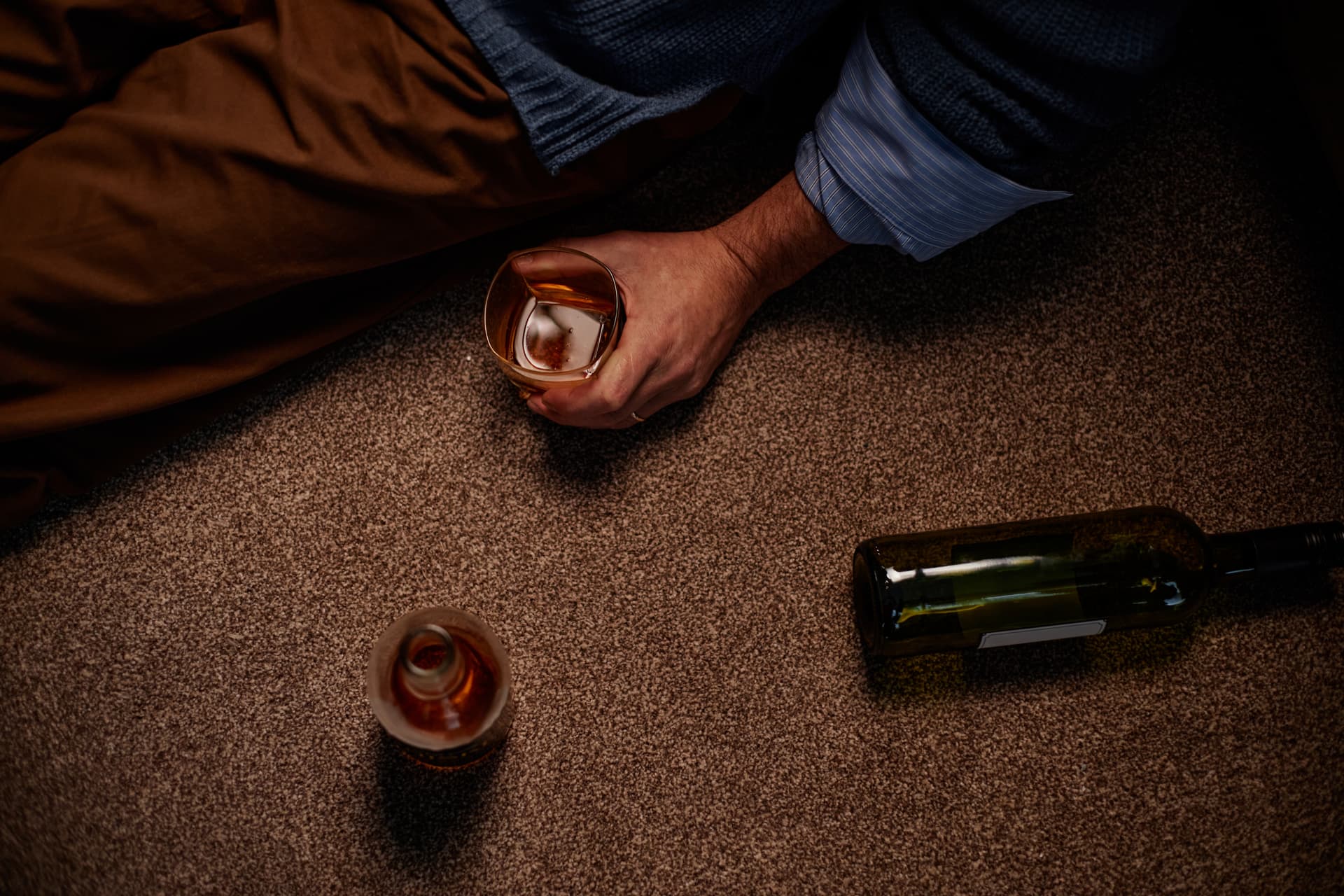 adult children of alcoholics — close-up of an adult sitting on carpet holding a whiskey glass, with two empty bottles nearby, suggesting hidden drinking at home.